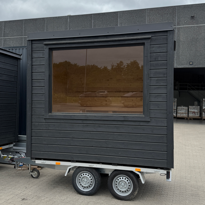 Black modular building on wheels with large window against a gray sky.